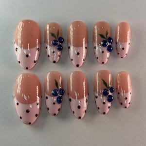 May include: A set of ten oval false nails. Each nail has a light pink base with black polka dots and a nude top. Some nails are decorated with blue berry and green leaf accents.