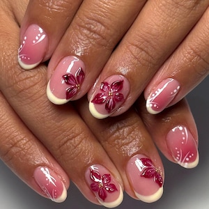 May include: Manicured nails with a pink and white gradient design. Each nail features a 3D burgundy flower with gold accents. The nails have a French tip design with a white crescent shape at the base.