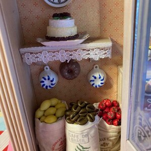 May include: A miniature dollhouse kitchen with a white cake with chocolate frosting, a clock, and three bags of goods. The bags are labeled "Cafe Do Brasil", "Australian", and "Mexican".