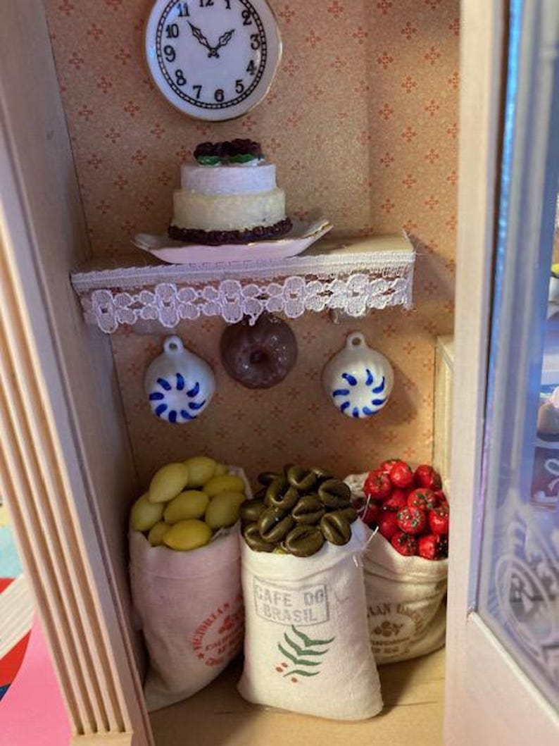 May include: A miniature dollhouse kitchen with a white cake with chocolate frosting, a clock, and three bags of goods. The bags are labeled "Cafe Do Brasil", "Australian", and "Mexican".