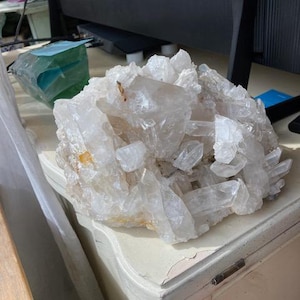 May include: A large cluster of clear quartz crystals, with some crystals having a slight yellow tint. The crystals are arranged in a natural, organic shape, and they are sitting on a white surface.