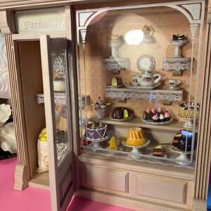 May include: A miniature wooden storefront with a glass display case filled with miniature pastries and cakes. The storefront is labeled "Patisserie" and has a door that is open.