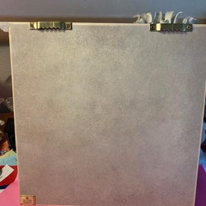 May include: The back of a light brown framed picture with two gold hanging hooks and a small gold label that says "Remarkable Designs".