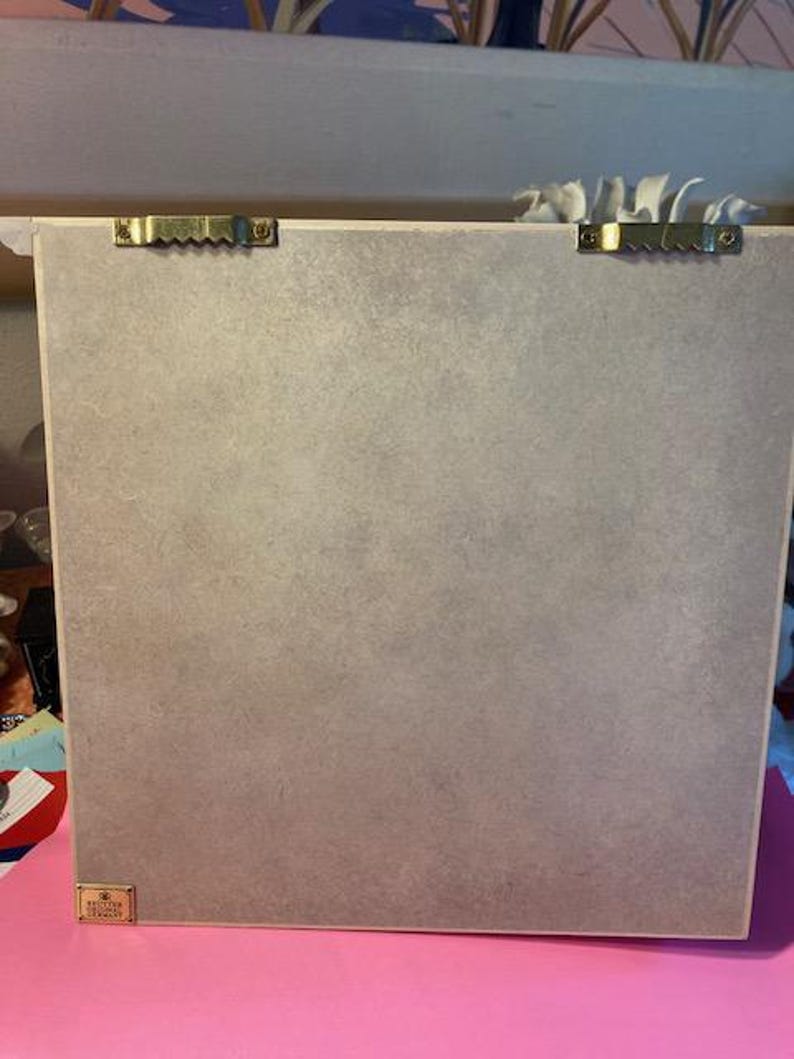 May include: The back of a light brown framed picture with two gold hanging hooks and a small gold label that says "Remarkable Designs".