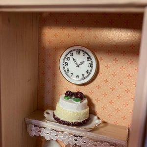 May include: A miniature dollhouse kitchen with a white cake with red roses on top, a clock on the wall, and a lace trim shelf.