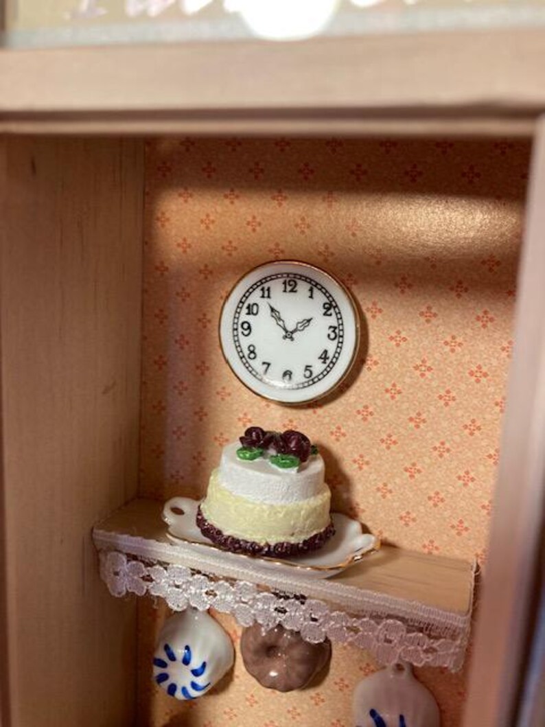 May include: A miniature dollhouse kitchen with a white cake with red roses on top, a clock on the wall, and a lace trim shelf.