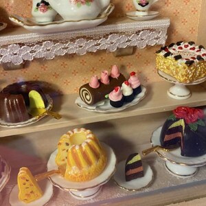 May include: A miniature dollhouse bakery display with various pastries and cakes, including a chocolate cake, a yellow cake with a white glaze, a chocolate roll with pink frosting, and a chocolate cake with a red rose on top.