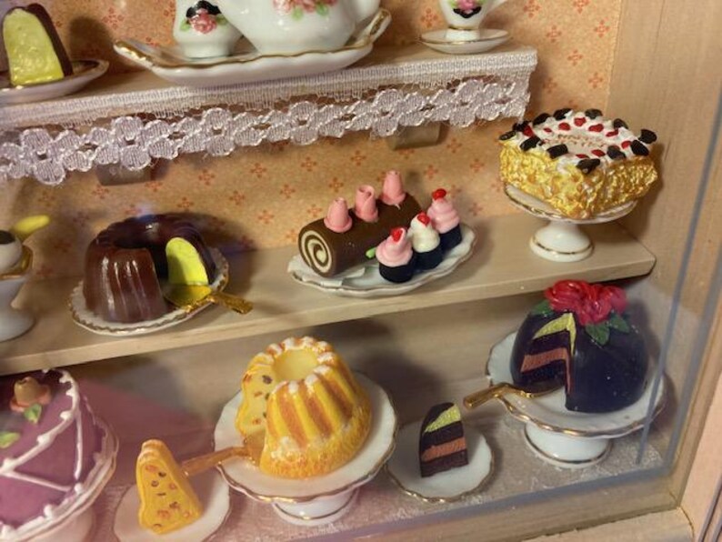 May include: A miniature dollhouse bakery display with various pastries and cakes, including a chocolate cake, a yellow cake with a white glaze, a chocolate roll with pink frosting, and a chocolate cake with a red rose on top.