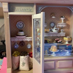 May include: A miniature wooden bakery shop with a glass display case filled with pastries. The shop has a sign that reads "Patisserie" and a clock on the wall. The display case is filled with cakes, cookies, and other treats.