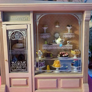 May include: A wooden miniature bakery display case with glass shelves filled with miniature cakes, pastries, and other treats. The case has a sign that reads "Patisserie".