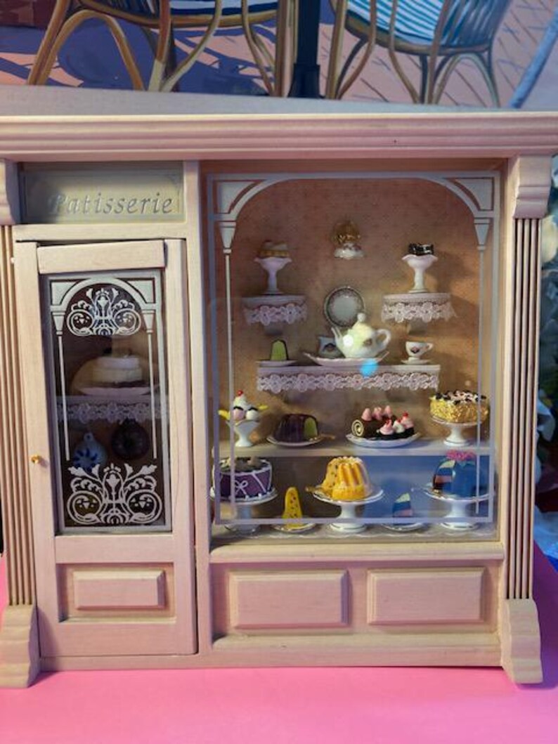 May include: A wooden miniature bakery display case with glass shelves filled with miniature cakes, pastries, and other treats. The case has a sign that reads "Patisserie".