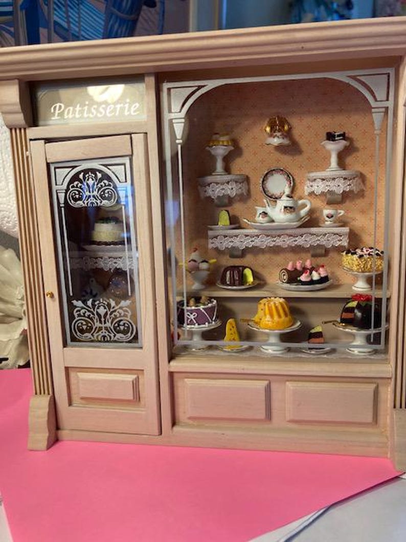 May include: A miniature wooden storefront with a glass display case filled with miniature pastries and cakes. The storefront is painted a light wood color and has a sign that reads "Patisserie".