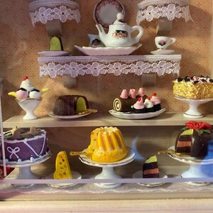 May include: A miniature bakery display case with a variety of pastries and desserts, including a chocolate cake, a bundt cake, a chocolate eclair, a strawberry cake, and a chocolate cake with a red rose on top.
