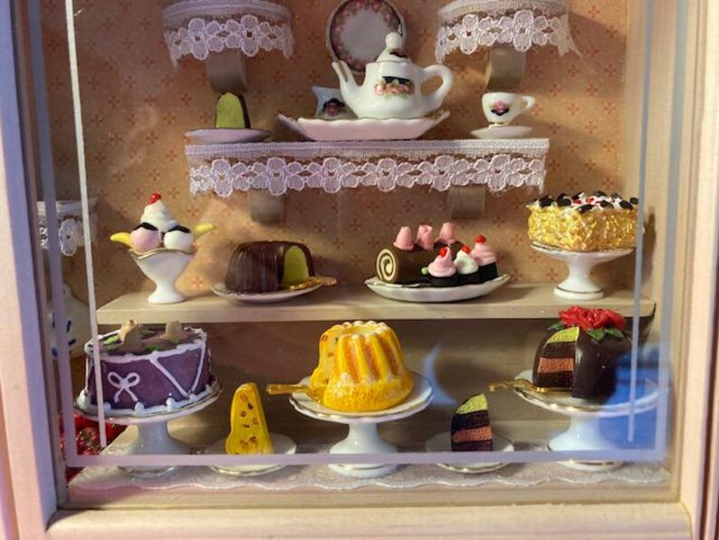 May include: A miniature bakery display case with a variety of pastries and desserts, including a chocolate cake, a bundt cake, a chocolate eclair, a strawberry cake, and a chocolate cake with a red rose on top.
