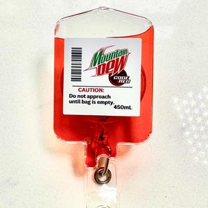 May include: A novelty badge reel shaped like a blood bag, filled with red liquid. The label reads "Mountain Dew Code Red" with a barcode and the warning "CAUTION: Do not approach until bag is empty, 450mL." The reel has a clear plastic clip.
