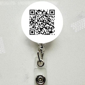 May include: A white, circular badge reel with a black and white QR code. The reel has a clear plastic strap with a snap closure and a silver-colored metal clip. The background is a light, speckled surface.