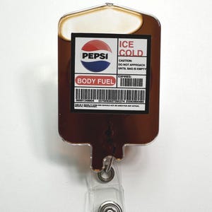 May include: A novelty Pepsi-themed badge reel shaped like an IV bag. The badge reel features the Pepsi logo, the words "BODY FUEL" and "ICE COLD", and a warning label. The reel has a clear plastic strap and a metal snap.