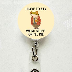 May include: A retractable badge reel with a light yellow circular design. The design features a cartoon frog in a suit holding a coffee cup, with the text "I HAVE TO SAY WEIRD STUFF OR I'LL DIE". The reel has a clear plastic strap and a metal clip.