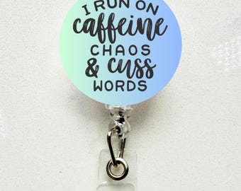 May include: A round badge reel with a gradient blue and green background. The badge reel has the text "I RUN ON caffeine CHAOS & cuss WORDS" in black script. The reel has a silver metal clip and a clear plastic holder.