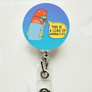 May include: A round badge reel with a cartoon fish character on a blue background. The fish is orange and wearing a light blue shirt. A yellow speech bubble says "THIS IS A LOAD OF BARNACLES". The reel has a silver clip and a clear plastic holder.