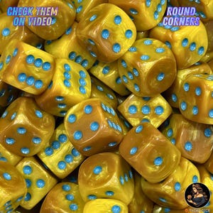 Puede incluir: Una colección de dados de seis caras con un acabado dorado nacarado y puntos de color azul brillante. Los dados tienen esquinas redondeadas. El texto en la imagen dice: CHECK THEM ON VIDEO y ROUND CORNERS. Los dados están en un montón denso.