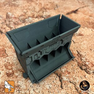 May include: A dark gray, rectangular storage container with a series of internal dividers. The container has a textured surface and a detailed, industrial design. The container is designed for organizing small items.