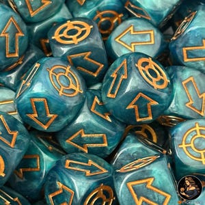 May include: A close-up shot of multiple teal-colored dice with gold-colored symbols and arrows. The dice have a marbled appearance. The symbols include arrows pointing up, down, left, and right, as well as a target symbol. The dice are for tabletop games.