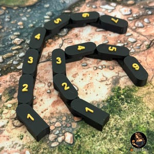 May include: Black, segmented game pieces with gold numbers from 1 to 9 arranged in a curved line. The pieces are on a textured, brown and green surface. The game pieces are likely for a board game or tabletop game.