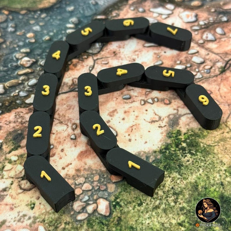 May include: Black, segmented game pieces with gold numbers from 1 to 9 arranged in a curved line. The pieces are on a textured, brown and green surface. The game pieces are likely for a board game or tabletop game.
