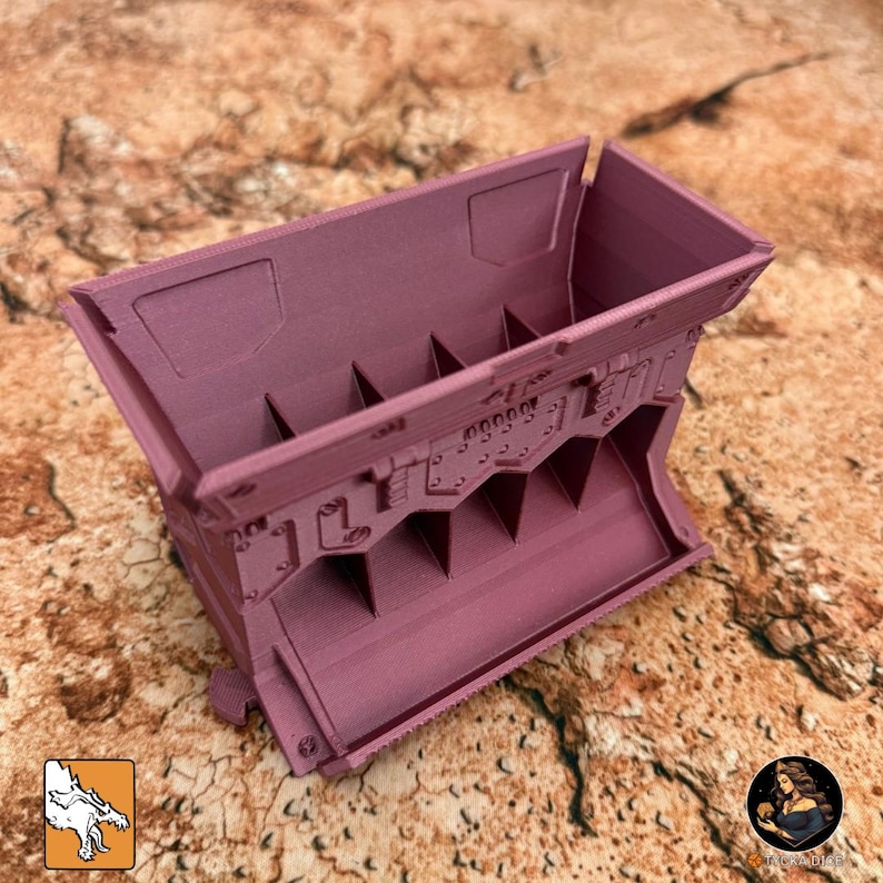 May include: A mauve-colored, rectangular container with internal dividers. The container has a detailed, industrial design with rivets and geometric shapes. The item is likely 3D-printed and sits on a textured, brown surface.