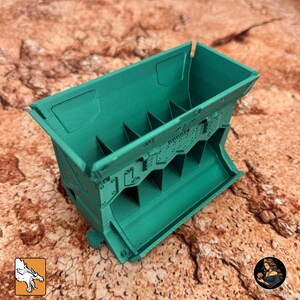 May include: A teal-colored, rectangular container with a series of internal dividers. The container has a detailed, textured surface with what appears to be a mechanical or industrial design. The object is likely a storage or organizational unit.