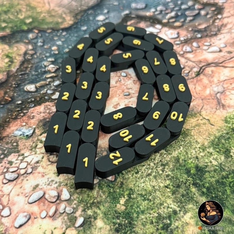 May include: Black, rectangular game pieces with gold numbers from 1 to 12 arranged in a chain. The pieces are on a textured, brown and green surface. The game pieces are likely for a board game or role-playing game.