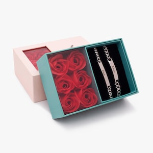 Puede incluir: Un juego de regalo con jabones en forma de rosas rojas y dos pulseras de cadena plateadas. Las pulseras están grabadas con nombres. La caja es turquesa y rosa, con una tapa transparente.