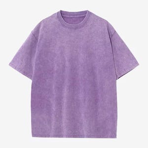 Może przedstawiać: T-shirt z kr&oacute;tkim rękawem i okrągłym dekoltem w jednolitym, wyblakłym fioletowym kolorze. Koszulka ma luźny kr&oacute;j i lekko teksturowany wygląd, sugerujący efekt vintage lub prania. Tkanina wydaje się miękka i wygodna.