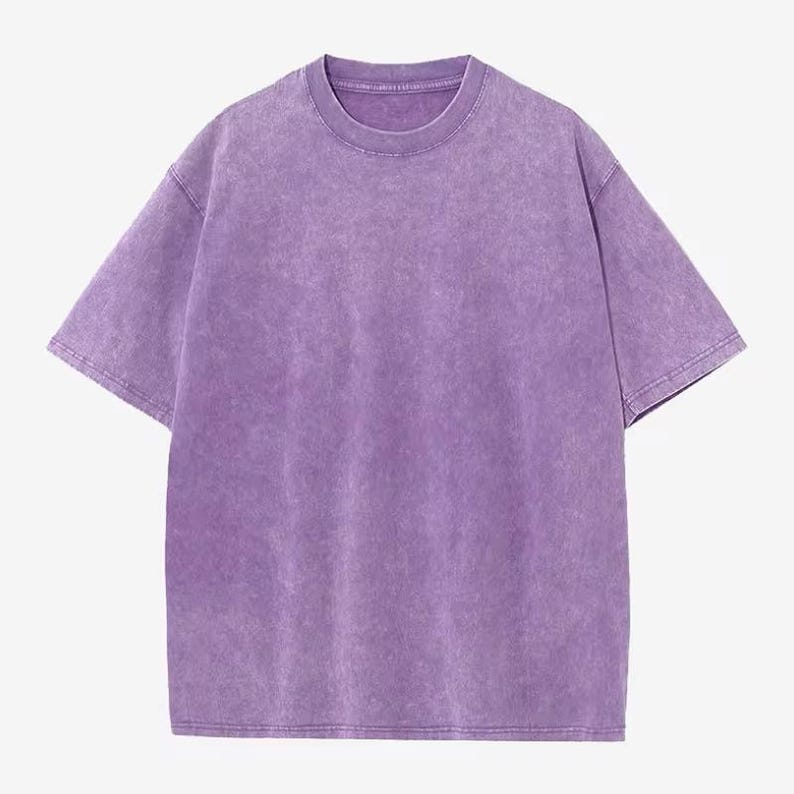 Może przedstawiać: T-shirt z kr&oacute;tkim rękawem i okrągłym dekoltem w jednolitym, wyblakłym fioletowym kolorze. Koszulka ma luźny kr&oacute;j i lekko teksturowany wygląd, sugerujący efekt vintage lub prania. Tkanina wydaje się miękka i wygodna.