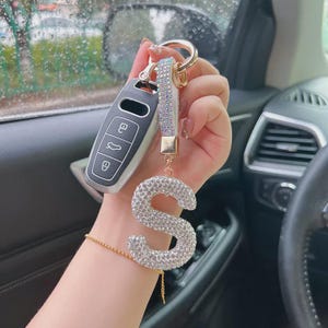 May include: A silver car key fob with a rhinestone-covered letter "S" keychain. The keychain has a gold-colored ring and a strap adorned with iridescent rhinestones. The background shows the interior of a car.