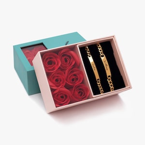 May include: A gift box with red roses and two gold bracelets. The bracelets have the names "Sophia" and "William" engraved on them. The box is light pink and teal, with a clear window to see the roses.