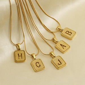以下が含まれることがあります： ゴールドカラーのネックレスで、それぞれ異なる文字（H、C、J、A、B）が表示された正方形のペンダントが付いています。ペンダントはテクスチャ加工された表面で、繊細なチェーンから吊り下げられています。背景は明るくニュートラルな色です。