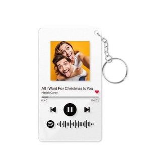 May include: A white keychain featuring a photo of a couple making silly faces. The design mimics a music streaming interface, displaying the song "All I Want For Christmas Is You" by Mariah Carey, with a play button and a sound wave graphic.