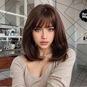 Op de afbeelding: Een vrouw met bruin haar en pony, draagt een gouden ketting en een beige top. De afbeelding toont het Aura Wigs-logo. De achtergrond is een keuken met een geometrisch patroon.