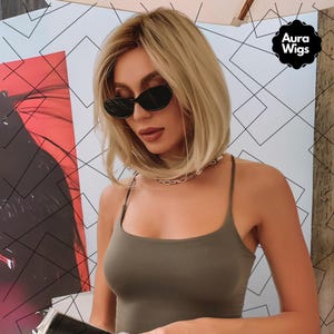 Puede incluir: Una mujer con cabello rubio que lleva un top verde sin mangas y gafas de sol negras. El fondo es un patrón geométrico con el texto "Aura Wigs" en un círculo.