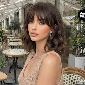 May include: A woman with wavy, dark brown hair and bangs. She is wearing a beige top and delicate jewelry. The image includes the Aura Wigs logo in the upper right corner.