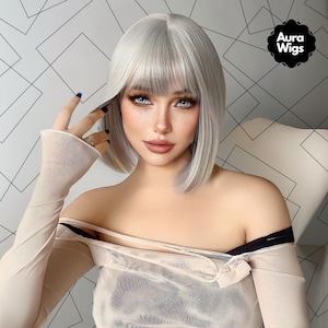 Puede incluir: Una peluca corta, recta, de color blanco plateado con flequillo recto. La peluca está peinada en un bob con raya lateral. La peluca la lleva una persona de piel clara y pecas.