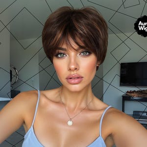 Puede incluir: Una mujer con una peluca marrón corta y en capas con reflejos y flequillo. Lleva una camiseta azul claro y un collar dorado con un colgante. El logotipo de Aura Wigs es visible en la esquina superior derecha.