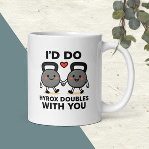 May include: White ceramic mug with the text "I'D DO HYROX DOUBLES WITH YOU". The mug features a design of two cartoon kettlebells holding hands with a heart above them. The mug has a large handle and is set against a neutral background.
