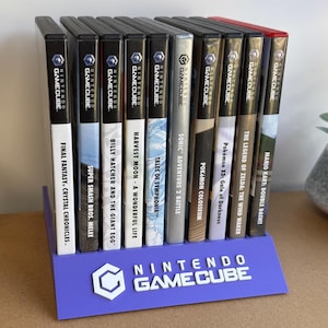 May include: A purple plastic display stand featuring the white Nintendo GameCube logo and text, holding a row of ten Nintendo GameCube video game cases. The cases are arranged vertically, showcasing titles such as Final Fantasy, Super Smash Bros, and Mario Kart.