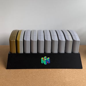 Könnte beinhalten: Ein schwarzer Kunststoff-Präsentationsständer mit einem bunten N64-Logo auf der Vorderseite, der zehn Videospielkassetten hält. Die Kassetten sind in einer Reihe angeordnet, mit einer goldenen Kassette links und neun grauen Kassetten daneben.