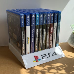 Peut inclure: Un support de présentation en plastique blanc contenant dix boîtiers de jeux PS4 alignés. Le support présente un logo PS4 noir en relief et un symbole PlayStation coloré sur le devant. Les titres incluent Uncharted 4, Ratchet & Clank, Call of Duty, Horizon Zero Dawn, Grand Theft Auto, Minecraft, Spider-Man, Rayman Legends et God of War.