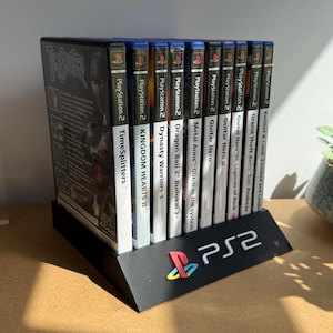 Könnte beinhalten: Ein schwarzer Kunststoff-Präsentationsständer mit neun PlayStation 2-Videospielhüllen in einer Reihe. Der Ständer zeigt das PS2-Logo in Weiß mit einem bunten PlayStation-Symbol. Die Spieltitel umfassen TimeSplitters, Kingdom Hearts II und weitere.