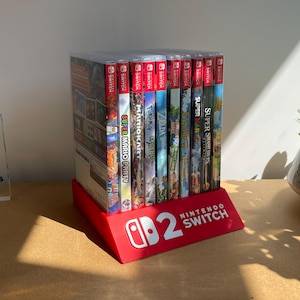 Könnte beinhalten: Ein roter Kunststoffständer zur Aufbewahrung von zehn Nintendo Switch-Spielehüllen, die in einer Reihe stehen. Auf der Vorderseite befindet sich das weiße Nintendo Switch-Logo und die Zahl zwei. Links steht ein transparenter Acrylblock mit der Aufschrift DISCONEXT MODS.
